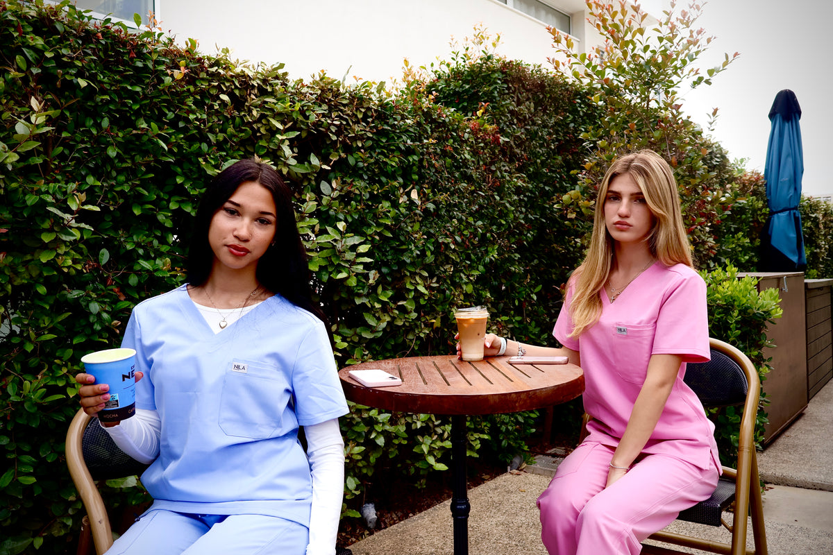 Two women wearing Nila scrubs, one in blush pink and one in light blue, in a professional setting